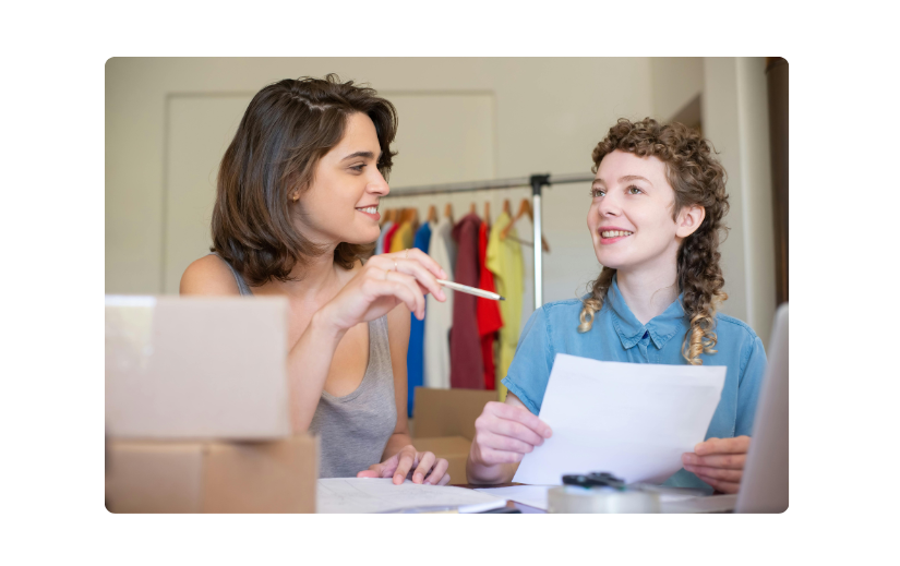 Two women planning an online store for beginners, with shipping boxes and clothing inventory in the background.